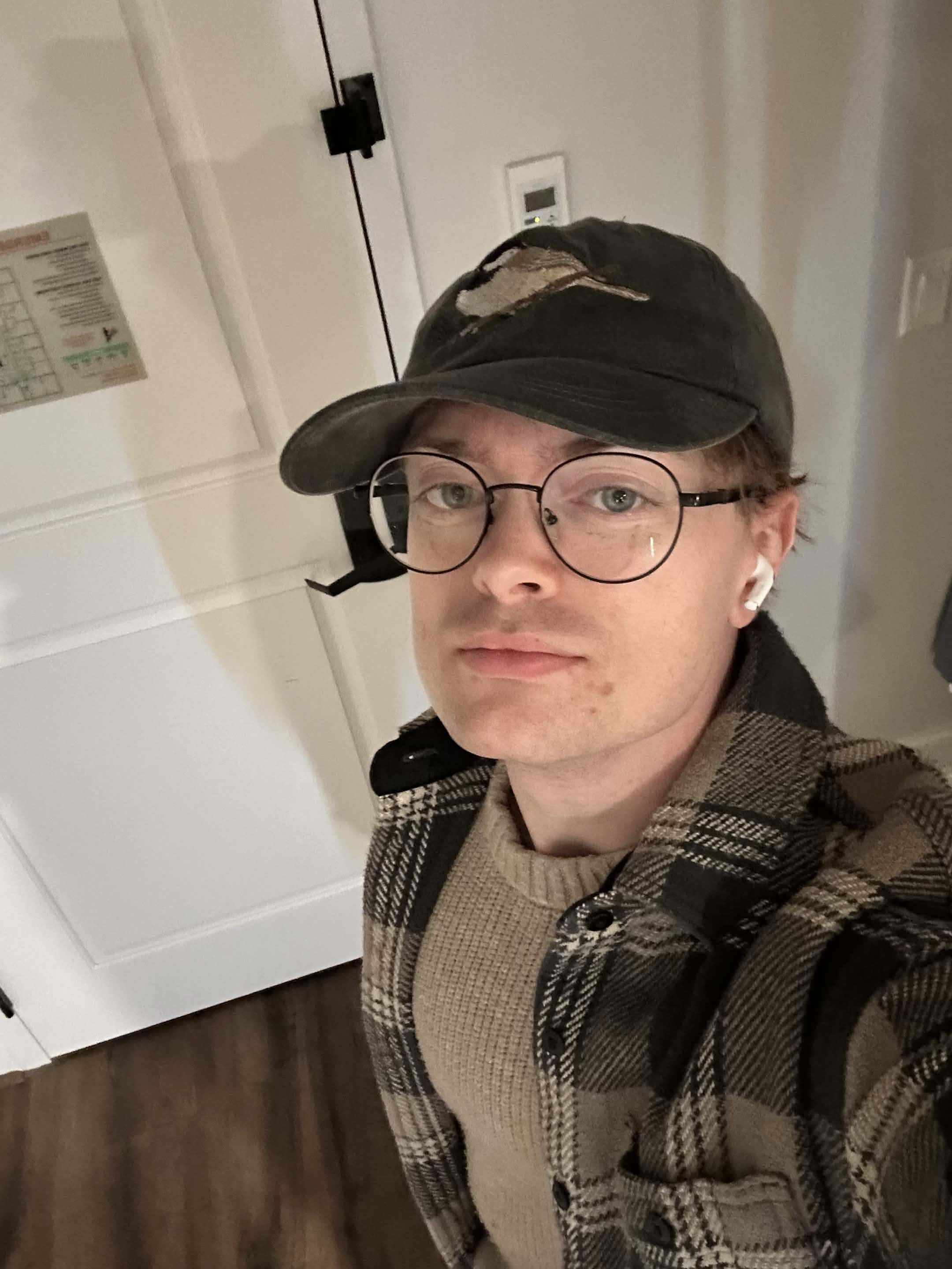 Photo of Jacob Hawkins wearing a cap, round glasses, and a plaid shirt.