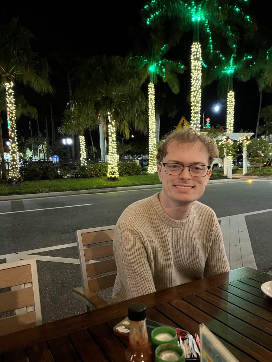 Jacob smiling at an outdoor restaurant table at night, with palm trees wrapped in holiday lights behind him.