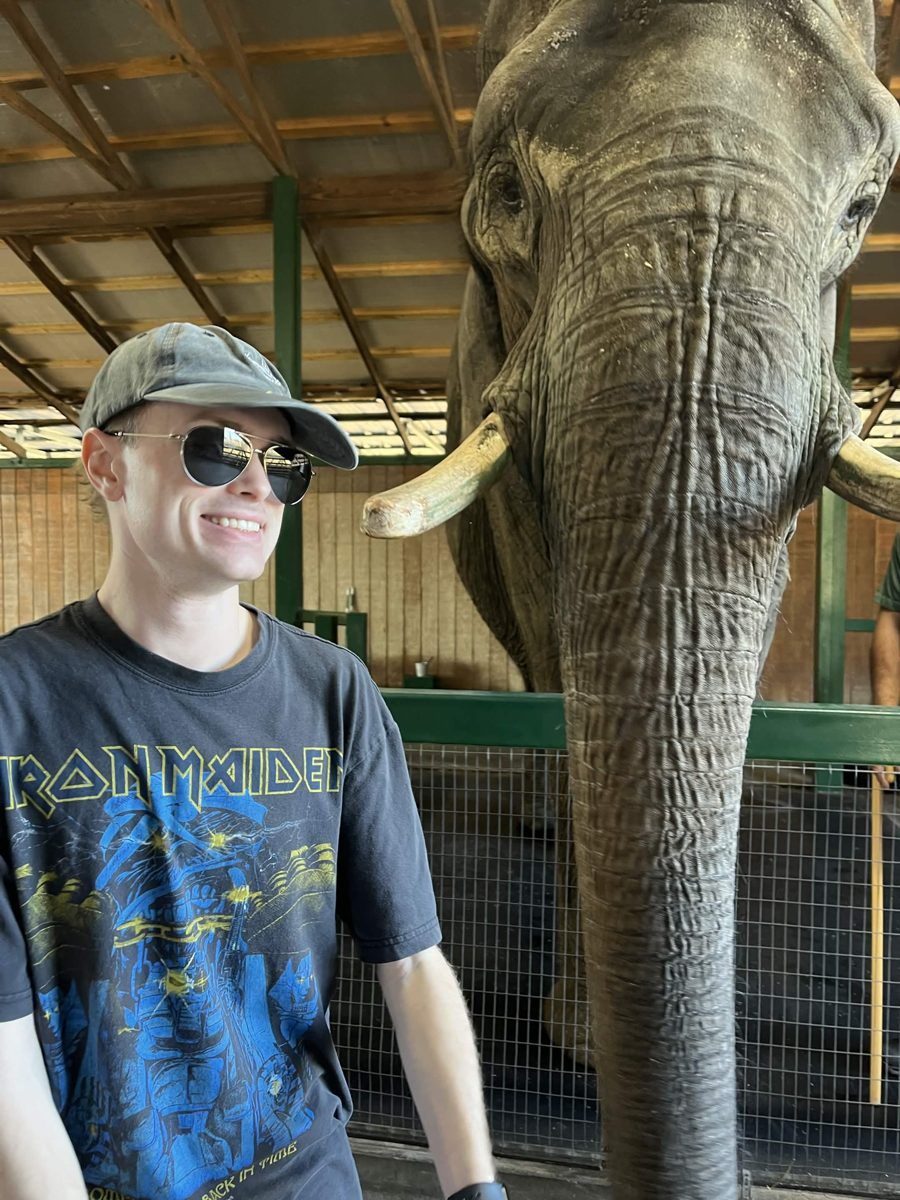Jacob in sunglasses and a cap, smiling next to an elephant.